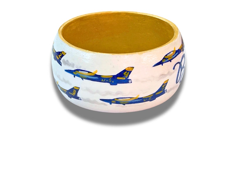 May include: A hand-painted wooden bangle bracelet with a white exterior and gold interior. The bracelet features blue and yellow jets with the text "Blue" in cursive. The jets are painted on a white background with gray clouds.