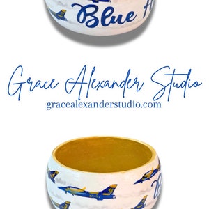May include: Hand-painted wooden bowl with a gold interior and white exterior. The exterior features blue jets and the words "Blue Angels" in blue script. The bowl is part of the "Grace Alexander Studio" collection.