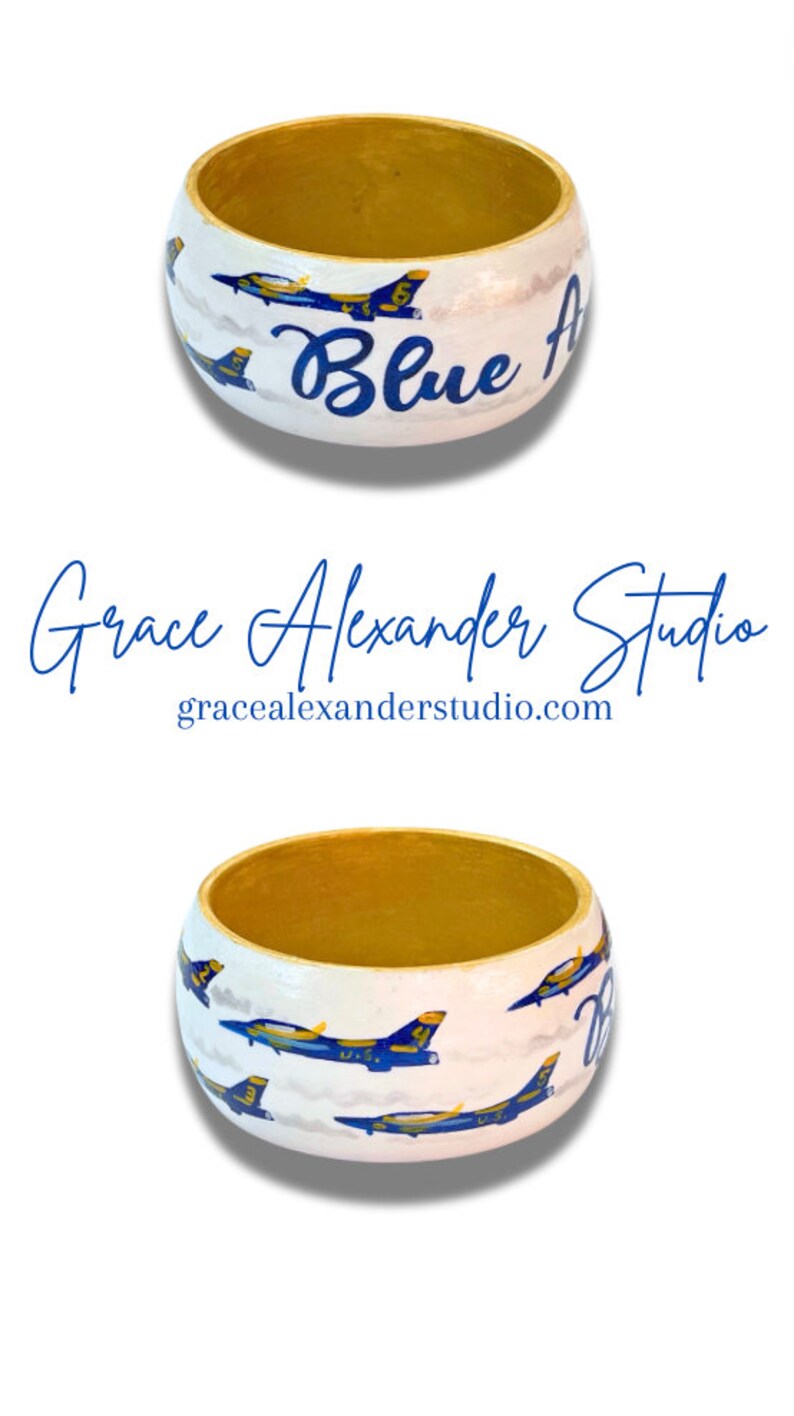 May include: Hand-painted wooden bowl with a gold interior and white exterior. The exterior features blue jets and the words "Blue Angels" in blue script. The bowl is part of the "Grace Alexander Studio" collection.