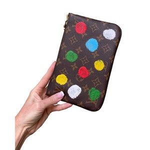 May include: Brown wallet with a gold zip, decorated with colourful polka dots in red, blue, green, yellow, and white. The wallet is held by a hand against a white background.