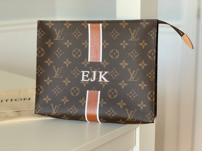 May include: Brown monogram clutch bag with white and brown stripes and the embroidered initials "EJK". The bag has a zip closure and a leather pull tab. The background is a white wall and a white shelf.