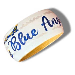 May include: A white wooden bangle bracelet with the words "Blue Angels" in blue script. The bracelet features a blue and gold airplane design and a gold interior.