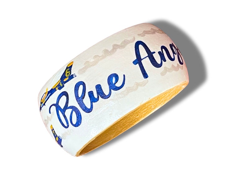 May include: A white wooden bangle bracelet with the words "Blue Angels" in blue script. The bracelet features a blue and gold airplane design and a gold interior.