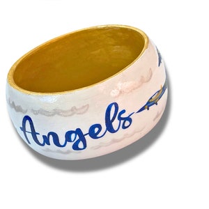 May include: A white, oval-shaped decorative bowl with a gold interior. The exterior features blue text that reads "Angels", along with painted clouds and blue and yellow airplanes. A unique, handcrafted item.