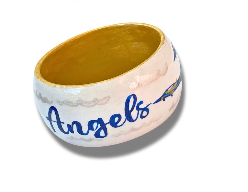May include: A white, oval-shaped decorative bowl with a gold interior. The exterior features blue text that reads "Angels", along with painted clouds and blue and yellow airplanes. A unique, handcrafted item.
