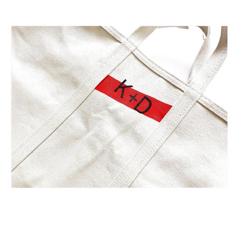 May include: Cream canvas tote bag with strong handles. A red rectangular patch is centered on the front, displaying the handwritten initials "K+D" in black. The bag has reinforced stitching along the seams and handles.