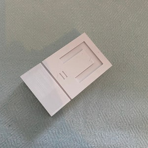 May include: A white plastic mold with a rectangular opening and two smaller rectangular openings. The mold is designed for creating a small object.