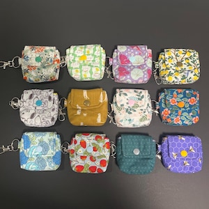 Peut inclure: Douze petites pochettes en tissu avec des fermoirs mousquetons en métal disposées sur trois rangées. Chaque pochette présente un rabat avec une fermeture à bouton-pression et divers motifs floraux, fruitiers, animaliers et géométriques.