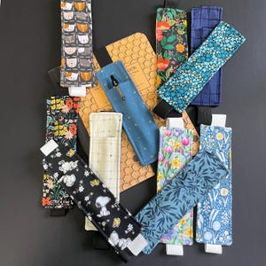 Peut inclure: Une collection de porte-stylos et de marque-pages en tissu avec divers motifs. Les motifs comprennent des visages de chats, des imprimés floraux, Snoopy et des formes géométriques. Chacun a un élastique noir et une extrémité blanche ou crème.