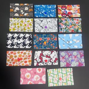 May include: A collection of fourteen rectangular fabric pouches with envelope-style flaps and snap closures. Each pouch features a distinct pattern, including cartoon characters, floral prints, geometric shapes, and animal motifs in various colours.