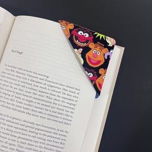 May include: A triangular corner bookmark featuring a black fabric background with a repeating pattern of colourful Muppet characters, including Fozzie Bear and Animal, placed over the top corner of an open book page with printed text.
