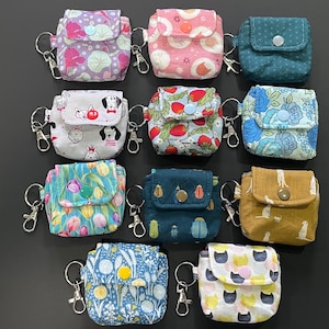 May include: A collection of eleven small fabric pouches with keychains, each featuring a flap closure secured by a plastic snap button. The pouches display various patterns including floral, animal, insect, and geometric prints in diverse colours.