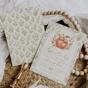 Pode incluir: Um cartão de convite para chá de bebê com uma ilustração em aquarela de um ursinho de pelúcia em uma maçã vermelha com um coração branco, cercado por folhagens. O texto diz The Apple of Our Eye Is On The Way, Grace and Jack, Oct 04.