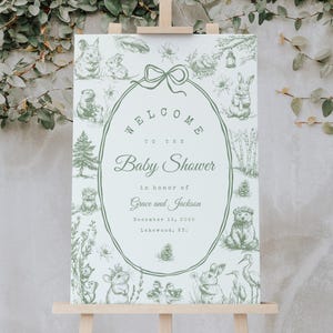 May include: A baby shower sign on a wooden easel. The sign has a green and white design with the words "Welcome to the Baby Shower" and the names "Grace and Jackson". The sign is surrounded by illustrations of animals.