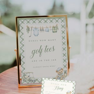 May include: A framed sign and a smaller card with a green and white plaid pattern. The sign reads "GUESS HOW MANY golf tees ARE IN THE JAR. CLOSEST GUESS WINS!" with illustrations of golf-themed baby clothes and a teddy bear.