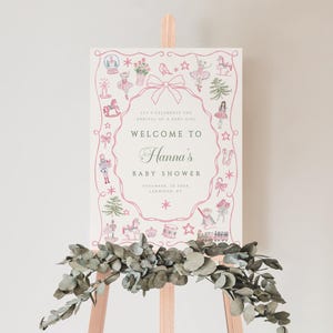 Könnte beinhalten: Ein Willkommensschild für eine Babyparty auf einer Holzstaffelei. Das Schild ist weiß mit einem rosa Rand und zeigt Illustrationen von Spielzeug und Balletttänzerinnen. Der Text lautet "Willkommen zu Hannas Babyparty" mit Datum und Ort.