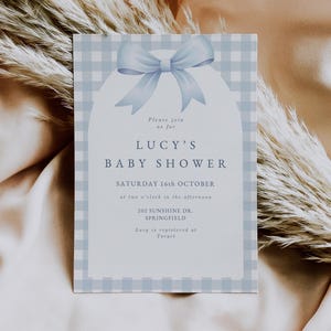 May include: A blue and white gingham baby shower invitation with a blue bow. The invitation reads "Please join us for Lucy's Baby Shower, Saturday 16th October at two o'clock in the afternoon, 202 Sunshine Dr., Springfield. Lucy is registered at Target."
