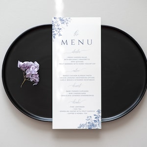 May include: A white menu card with blue floral illustrations and text that reads "the MENU" and lists various food and drink options. The card is on a black plate with a small purple flower.