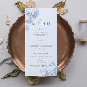 May include: A white menu card with blue and gray illustrations of hot air balloons. The menu card lists the food and drinks served at a wedding reception. The text on the menu card reads "The Menu", "Starters", "Fresh Garden Salad with Balsamic Vinaigrette", "Mini Quiches", "Fruit Skewers with Honey Yogurt Dip", "Entree", "Baked Chicken Alfredo Pasta", "Veggie Lasagna (Vegetarian Option)", "Garlic Bread & Steamed Vegetables", "Dessert", "Baby Themed Cupcakes", "Mini Cheesecakes", "Chocolate Covered Strawberries", "Drinks", "Pink Lemonade", "Iced Tea", "Sparkling Water with Fruit Garnish", "Coffee & Herbal Tea".