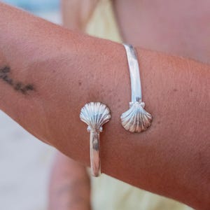 Può includere: Un bracciale a polsino argentato con un design a conchiglia. Il bracciale aperto presenta due ornamenti a forma di conchiglia, uno a ciascuna estremità. Il bracciale è indossato sul braccio, mettendo in risalto gli intricati dettagli degli abbellimenti a forma di conchiglia. Il gioiello è realizzato in metallo lucido e riflettente.