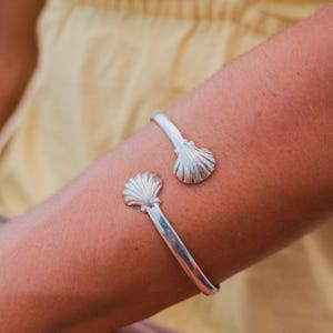 May include: A silver cuff bracelet featuring two identical shell-shaped terminals at each end. The metal has a polished, reflective finish, and the shell designs include fine, radiating grooves that extend from the base to the outer edge.