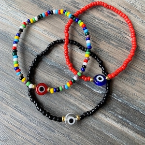 Op de afbeelding: Drie kralenarmbanden met boze oog-charms. Een armband is zwart met een witte boze oog-charm, een is rood met een blauwe boze oog-charm en een is veelkleurig met een rode boze oog-charm.
