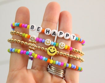 May include: A set of four colorful beaded bracelets with gold beads. The top bracelet features white beads with the words "BE HAPPY" in black lettering. The other bracelets feature colorful beads and smiley face charms.
