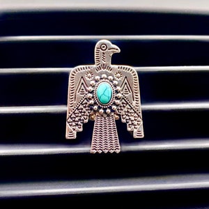 May include: A silver-toned metal car air freshener clip shaped like a stylized thunderbird. The bird features intricate geometric engravings on its wings and body, with a central oval-shaped turquoise stone set in a decorative beaded frame.