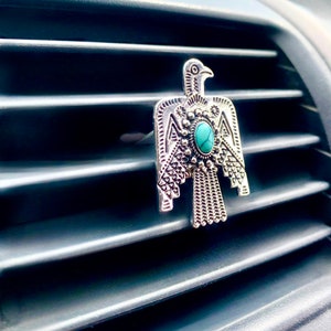 May include: A silver-toned metal car air vent clip shaped like a stylized bird with intricate embossed geometric patterns and a single oval turquoise-colored stone set in the center of the chest, attached to a horizontal vehicle air vent.