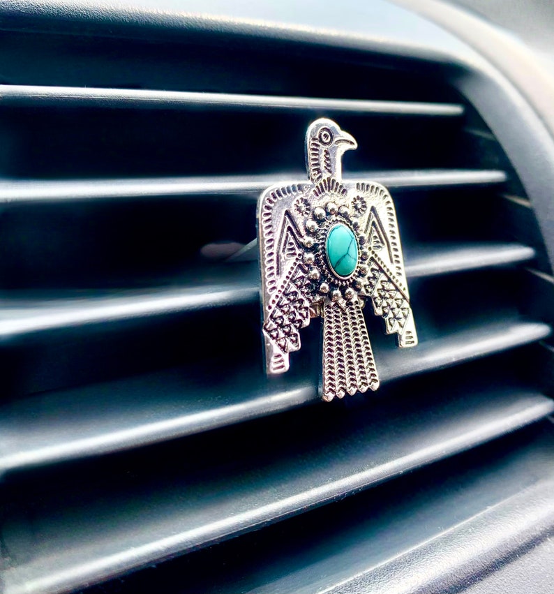 May include: A silver-toned metal car air vent clip shaped like a stylized bird with intricate embossed geometric patterns and a single oval turquoise-colored stone set in the center of the chest, attached to a horizontal vehicle air vent.