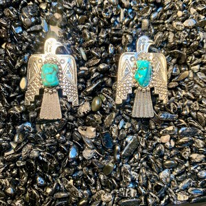May include: Two silver-toned metal pins shaped like eagles, each featuring a central rough-cut turquoise stone. The eagle designs include intricate geometric patterns on the wings and tail feathers, with a beaded border surrounding the turquoise.