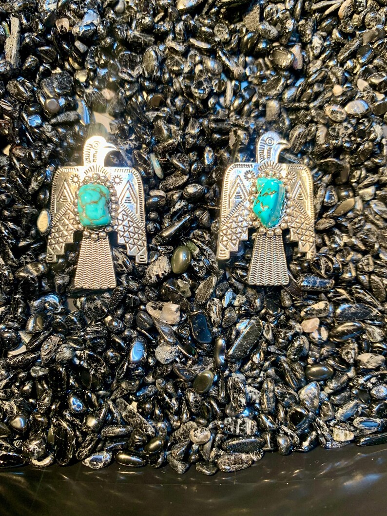 May include: Two silver-toned metal pins shaped like eagles, each featuring a central rough-cut turquoise stone. The eagle designs include intricate geometric patterns on the wings and tail feathers, with a beaded border surrounding the turquoise.