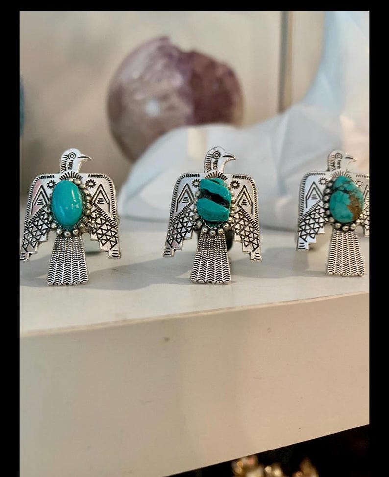 May include: Three silver-toned rings featuring a bird-shaped design with engraved wing patterns and a central turquoise stone. Each ring displays a unique natural turquoise cabochon, showcasing variations in color and matrix patterns on the bird.