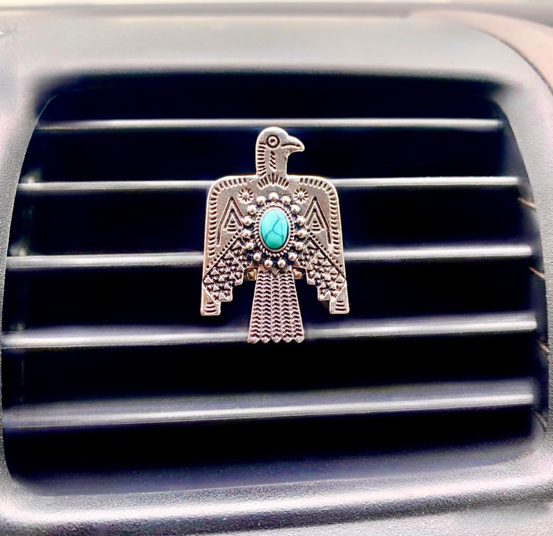 May include: A silver-toned metal car air freshener clip shaped like a stylized thunderbird. The bird features intricate geometric engravings on its wings and body, with a central oval-shaped turquoise stone set in a decorative beaded frame.