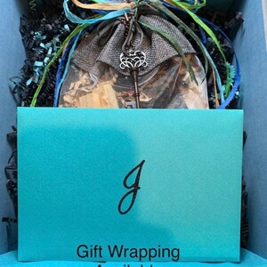 May include: A gift box containing a gray burlap drawstring pouch tied with blue and green ribbons, featuring a silver-toned ornate skeleton key charm. A turquoise card with a black letter J and the text Gift Wrapping Available sits in front.