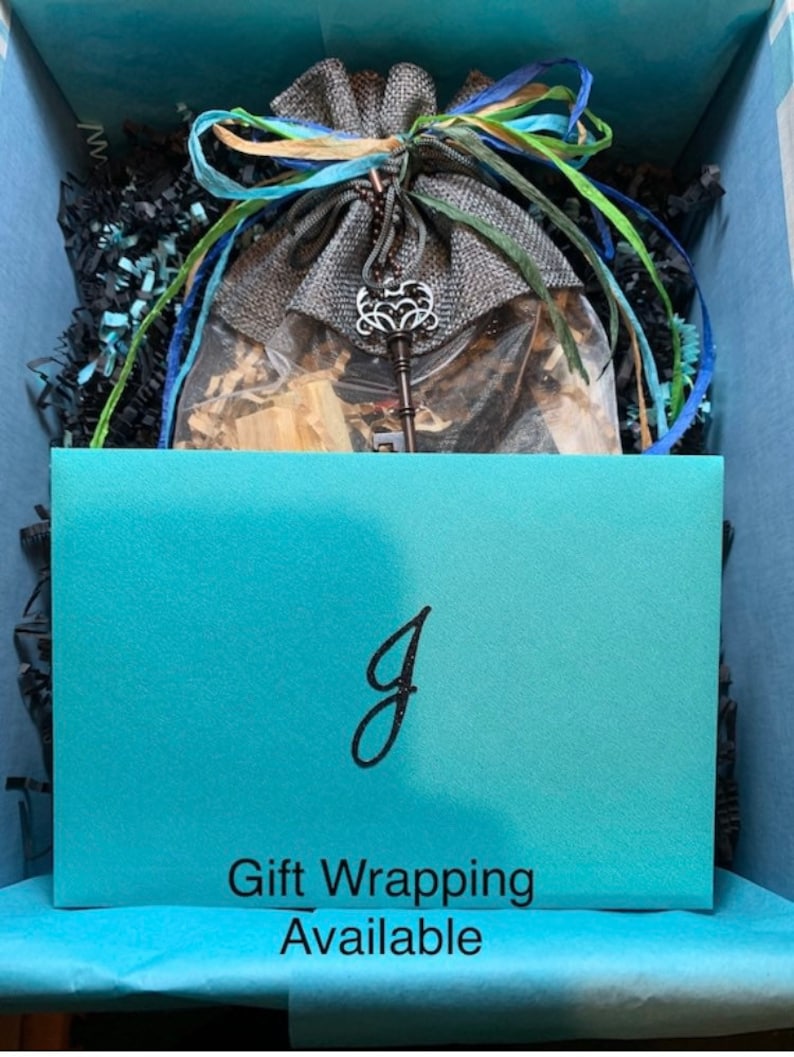 May include: A gift box containing a gray burlap drawstring pouch tied with blue and green ribbons, featuring a silver-toned ornate skeleton key charm. A turquoise card with a black letter J and the text Gift Wrapping Available sits in front.