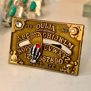 May include: A gold-toned Ouija board with a planchette in the shape of a hand with a red jewel. The board has the letters of the alphabet, numbers 1-9 and 0, and the words "yes", "no", "goodbye", and "Ouija Mystifying Oracle".
