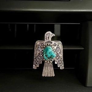 May include: A silver-toned metal brooch shaped like a bird with outstretched wings. The center of the bird features a rough-cut, bright turquoise stone surrounded by small silver beads. The metal surface is detailed with etched geometric patterns.