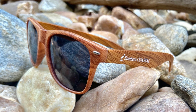 May include: Brown wood-framed sunglasses with dark lenses. The sunglasses have a white logo that says "Southern Coastal" with a bird design.