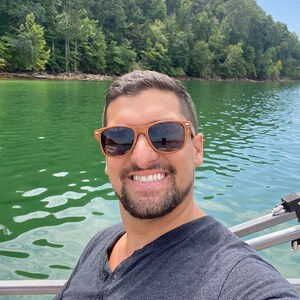 May include: A man wearing sunglasses and a grey t-shirt smiles at the camera while sitting on a boat on a lake. The lake is surrounded by green trees.