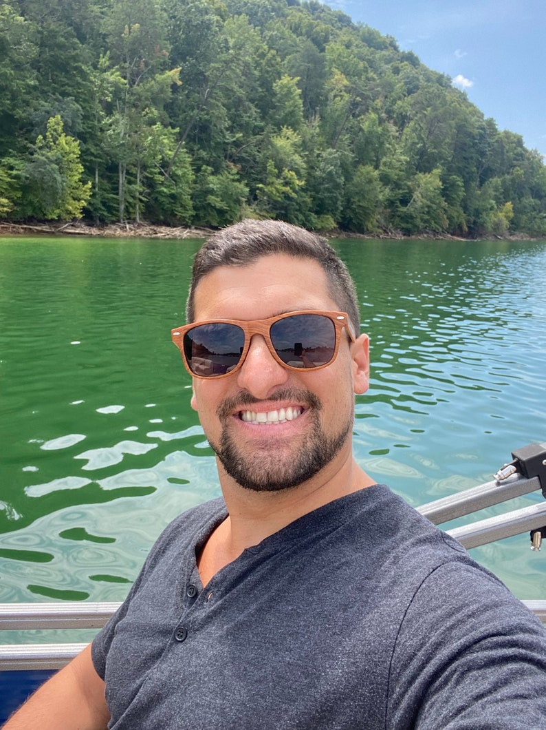 May include: A man wearing sunglasses and a grey t-shirt smiles at the camera while sitting on a boat on a lake. The lake is surrounded by green trees.