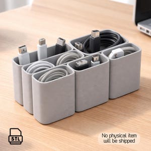 Pode incluir: Um organizador de cabos cinza com vários compartimentos, preenchido com vários cabos USB em branco e preto. O organizador está sobre uma superfície de madeira clara. O texto "STL" está no canto inferior esquerdo. O texto "No physical item will be shipped" está no canto inferior direito.