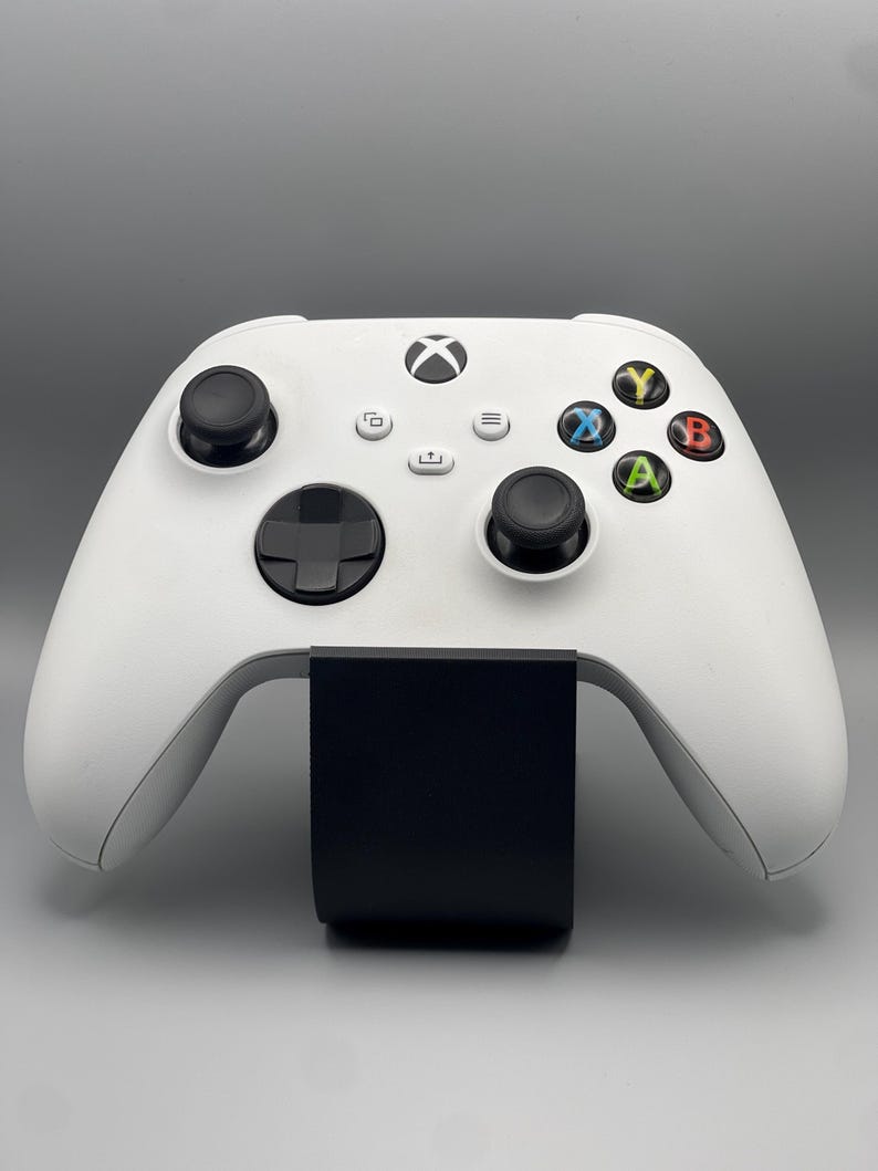 May include: A white Xbox Series X controller with black buttons and a black stand. The controller has a white Xbox logo on the front.