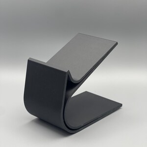 May include: A black, curved, plastic phone stand. The stand is designed to hold a phone upright.