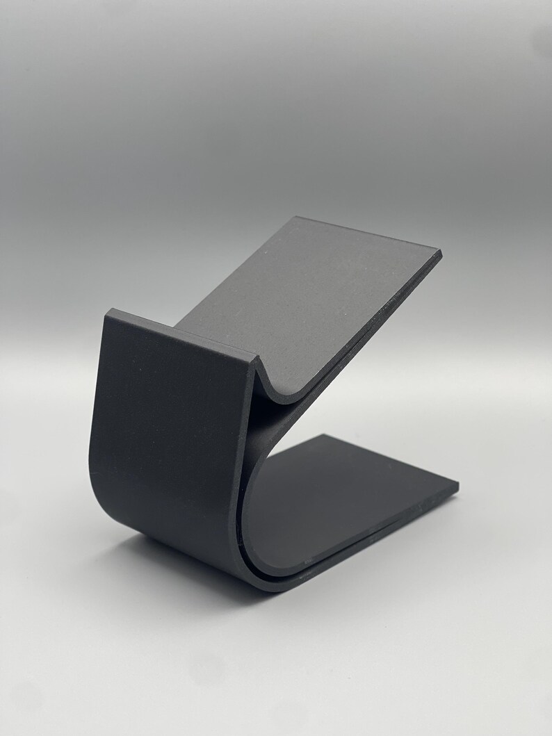 May include: A black, curved, plastic phone stand. The stand is designed to hold a phone upright.