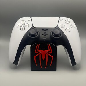 May include: A white PlayStation 5 DualSense controller with black accents and a red spider logo on a black stand.