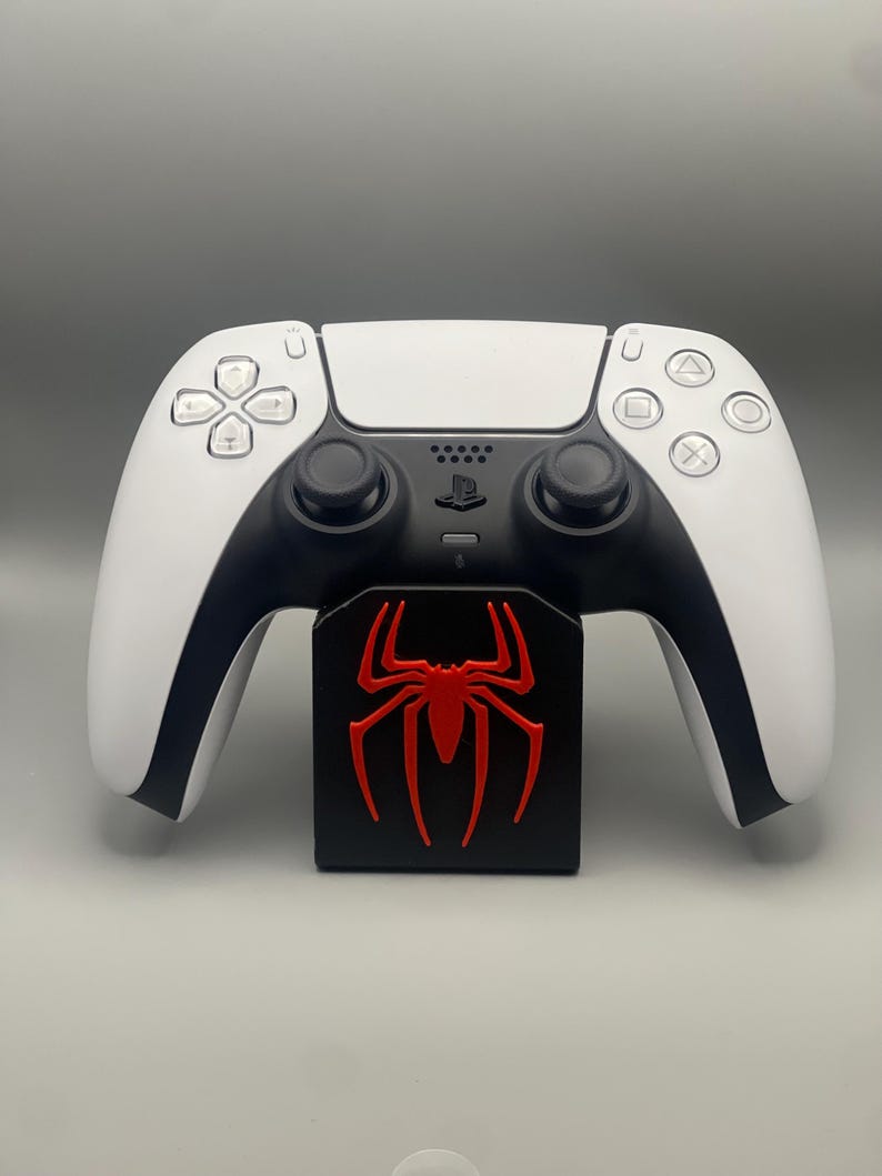 May include: A white PlayStation 5 DualSense controller with black accents and a red spider logo on a black stand.