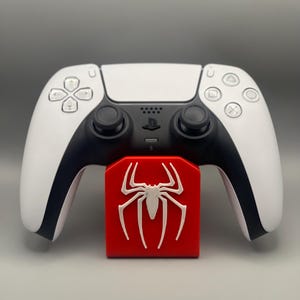 May include: A white PlayStation 5 DualSense controller is resting on a red stand with a white spider-man logo.
