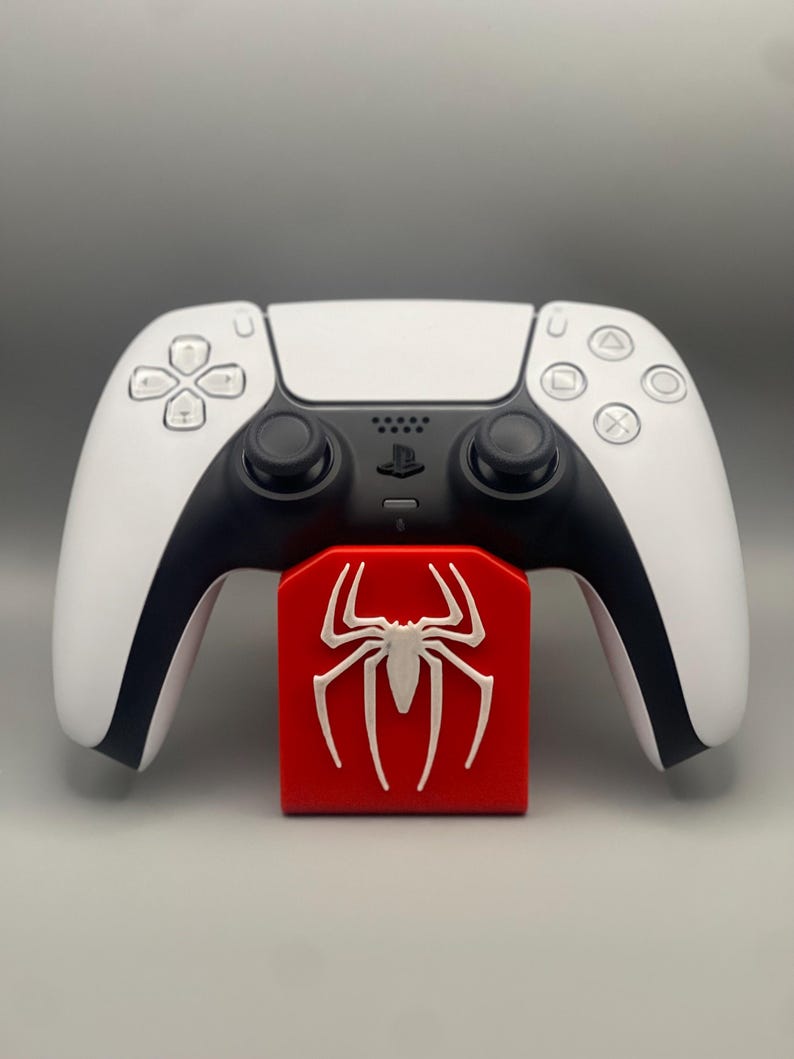 May include: A white PlayStation 5 DualSense controller is resting on a red stand with a white spider-man logo.