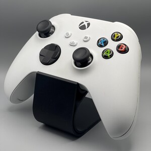 May include: A white Xbox Series X controller with black buttons and a black stand. The controller has a black Xbox logo on the front.